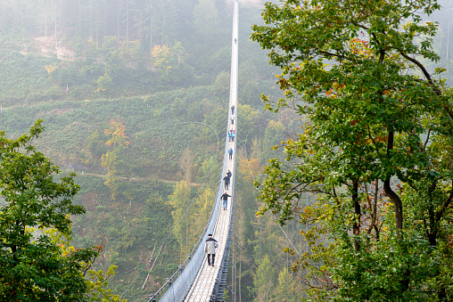 Suspension wooden bridge with steel ropes over a dense forest in West Germany, visible tourists on the bridge in misty weather.