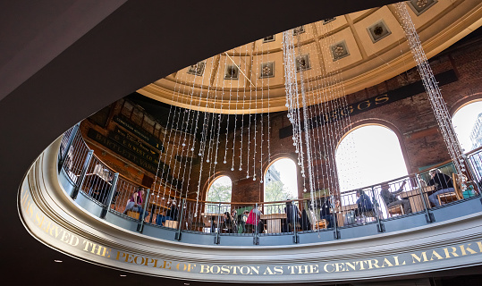Boston, Massachusetts, USA - October 22, 2021: Upward view of  Quincy Market building's rotunda with its second story open common seating area and copper-based dome above. Quincy Market (c. 1824-26) is a long two-story building behind Faneuil Hall in downtown Boston. It is a popular and busy lunchtime spot for downtown workers and tourist.  The sign boards of old businesses are seen on the brick walls.
