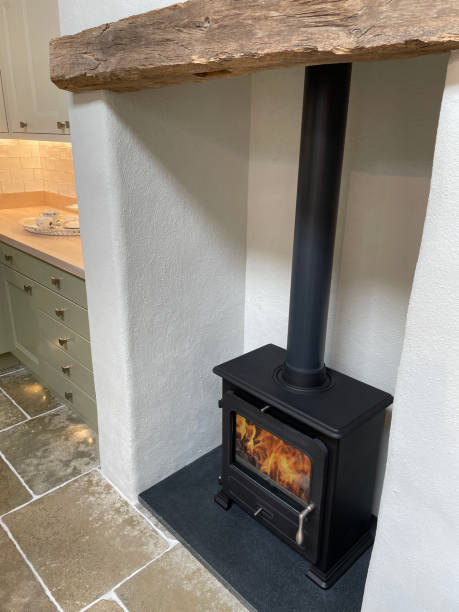 image d’un feu de gaz carré en fonte conçu comme un poêle à bois sur un foyer en ardoise dans une cuisine domestique, linteau à poutres en bois, flammes orange, tuyau de poêle en fonte et collier de fumée, plan de travail et armoires de cuisine - gas boiler water heater flame exploding photos et images de collection