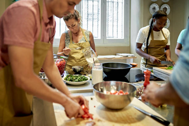 Adult Students Preparing Ingredients for Gazpacho Focus on background businesswomen in aprons slicing and seasoning ingredients for dish prepared during team cooking class. cooking class photos stock pictures, royalty-free photos & images