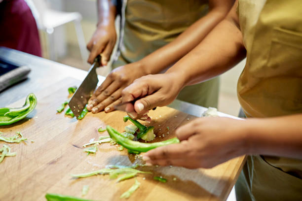 Adult Students Slicing Vegetable in Cooking Class Close-up of African and Colombian women in aprons standing at cutting board slicing green bell peppers. cooking class photos stock pictures, royalty-free photos & images