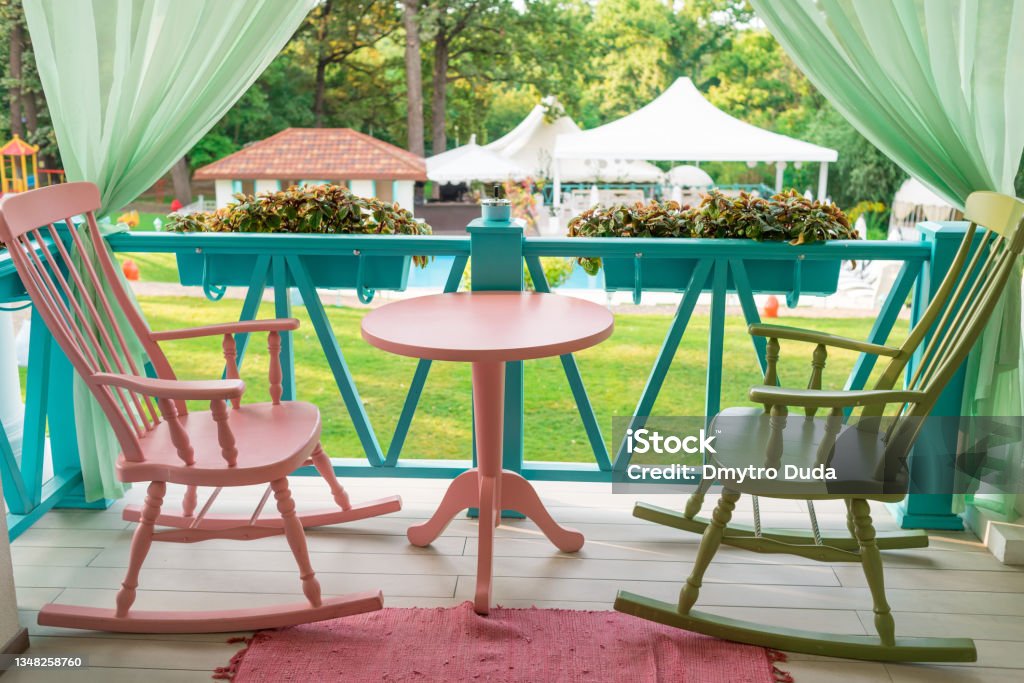 Pink and green wooden rocking chairs and table on porch or balcony. Two relaxing armchairs on porch of luxury hotel. Empty cafe with rocking chairs and table on summer  terrace outdoor, free space Multi Colored Stock Photo