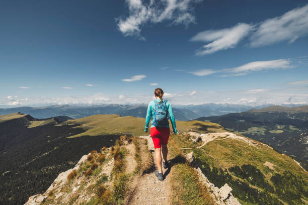 Back view of female hiker, backpacker walking on a narrow footpath on Seceda mountain in Dolomites, Italy.
