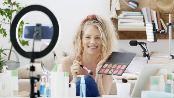 istock Blonde smiling woman influencer blogger recording a video tutorial on smartphone camera showing how to apply makeup cosmetics. Female vlogger making a beauty vlog for her online internet channel. 1348263038