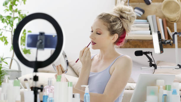 istock Blonde smiling woman influencer blogger recording a video tutorial on smartphone camera showing how to apply lips makeup cosmetics. Female vlogger making a beauty vlog for her online internet channel. 1348263091