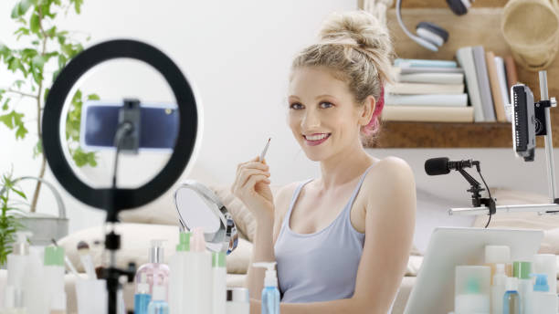 istock Blonde smiling woman influencer blogger recording a video tutorial on smartphone camera showing how to apply lips makeup cosmetics. Female vlogger making a beauty vlog for her online internet channel. 1348264771