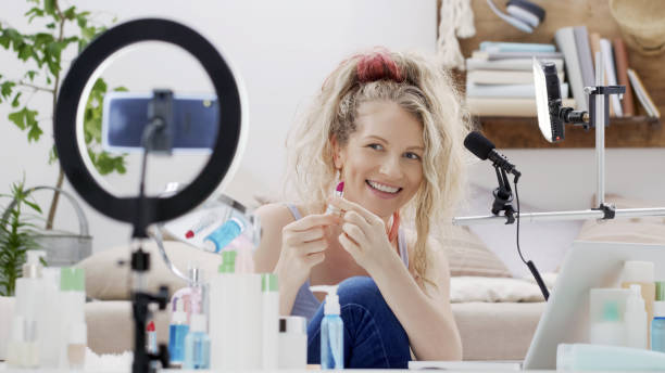 istock Blonde smiling woman influencer blogger recording a video tutorial on smartphone camera showing lipstick. Female vlogger making a beauty vlog for makeup products her online internet channel. 1348265301