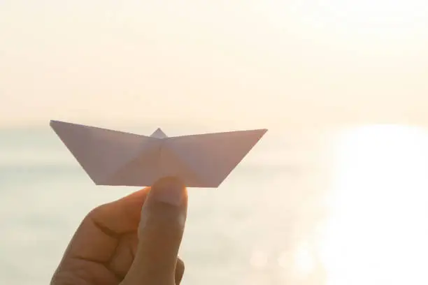 Hand of unrecognizable caucasian female at the sea  is showing a white paper boat at camera.