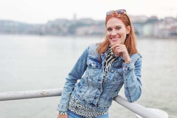 istock Young cheerful redhead woman looking at railing into camera at riverbank 1348288051