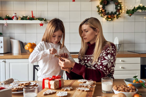 mamá ayudando a la linda hija pequeña a hacer galletas navideñas preparando regalos juntos sentados en la mesa de la cocina. madre e hija haciendo decoraciones familiares de pan de jengibre navideñas en casa. - christmas child cookie table fotografías e imágenes de stock