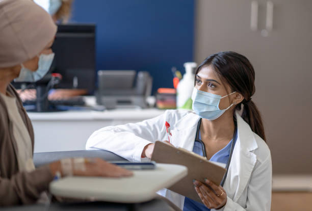 Doctor Talking with a Cancer Patient A female doctor bend's down to eye level as she talks with a cancer patient who is receiving her treatment intravenously. She is wearing a white lab coat and has a clipboard in hand as she takes notes. The patient is dressed casually and she sits back in the chair waiting for her treatment to finish, and both are wearing medical masks to protect them from COVID. medical research stock pictures, royalty-free photos & images