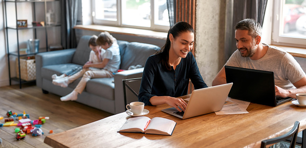 Wide banner view of happy young Caucasian parents couple sit at table at home work online on laptops. Smiling mother and father stay at home freelance on computer. Distant job, lockdown.