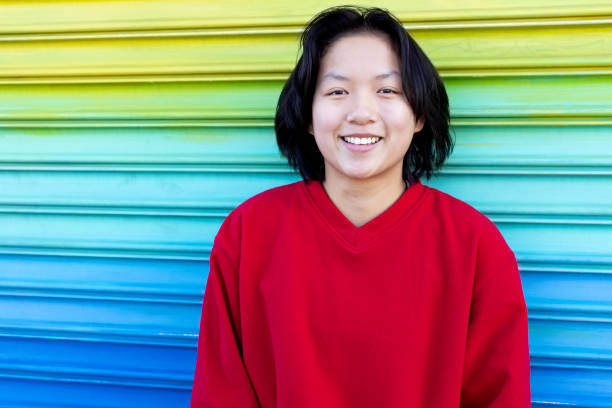 Close up  of smiling Asian teenager with short hair Androgynous female smiles in front of rainbow-colored wall outdoors androgyn stock pictures, royalty-free photos & images