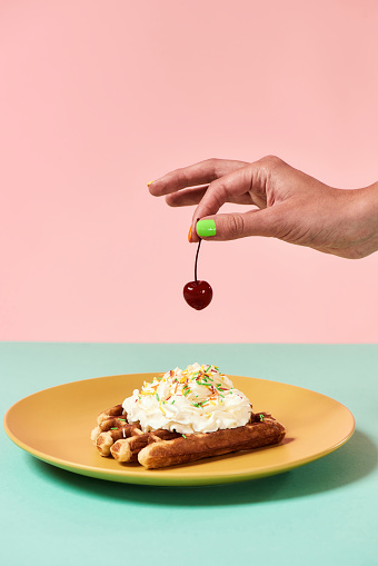 Close up of traditional Belgian waffles with whipped cream on a plate isolated over pastel pink green background. Hand holding and adding cherry on top. Vertical shot