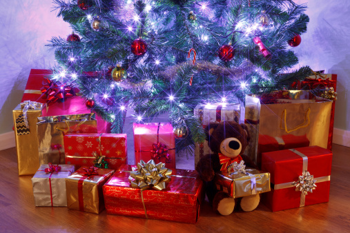 Photo of a Christmas tree with decorations and fairy lights surrounded by presents on a wooden floor. 