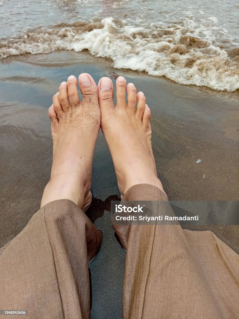 A Person's Feet On The Beach In Pangandaran, West Java, Indonesia. The legs of someone who is seen wearing brown trousers, is on the beach with the waves approaching him. This photo was taken on the West Coast, Pangandaran, West Java, Indonesia. Adult Stock Photo