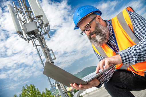Bearded technician in reflective vest working on laptop computer outdoors. Mobile signal antenna in background.