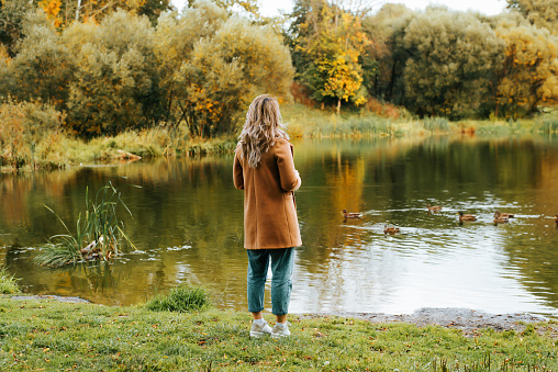 A tourist in a stylish sweater and hat with a backpack admires the view of the river. A young girl in a hat enjoys nature. Travel concept. Autumn mood.