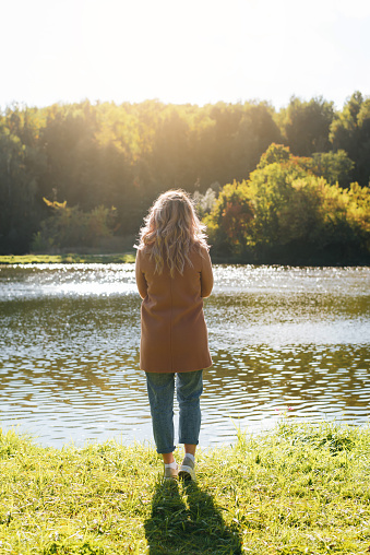 Back view of lonely young blonde woman in brown coat standing on shore near water against backdrop of lake and forest in afternoon outdoors. Contemplation, loneliness, enjoyment, freedom concept.