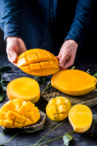 Men's hands hold a large juicy piece of yellow mango on a dark background