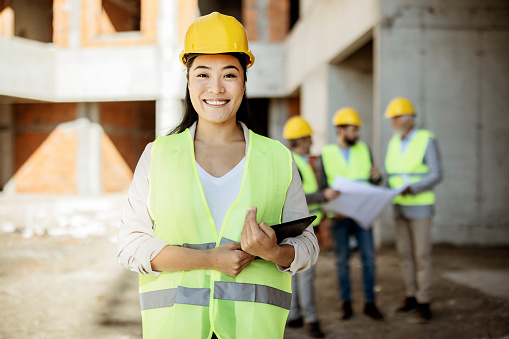 Portrait of an Asian Female Architect at Building Site Looking at Camera. Confident Construction Manager Wearing a Hardhat in Front of Her Colleagues