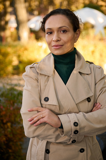 Confident portrait of attractive business woman in casual beige trench coat and green pullover looking at the camera, posing with crossed arms against the background of autumn nature in an urban park
