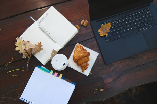 Flat lay autumnal composition with open notepad and copybook, laptop with fallen oak leaves, takeaway paper cup with hot drink and delicious baked croissant on a wooden table in the forest cafe