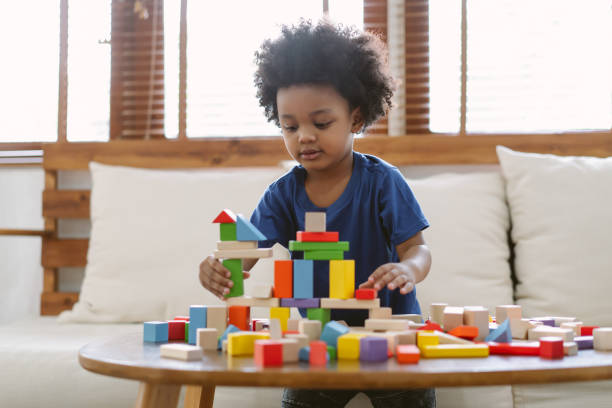 kleiner afroamerikanischer junge, der ein kleines haus mit bunten holzblöcken im wohnzimmer zu hause baut. lernspielzeug für vorschul- und kindergartenkinder. - kleinstkind stock-fotos und bilder