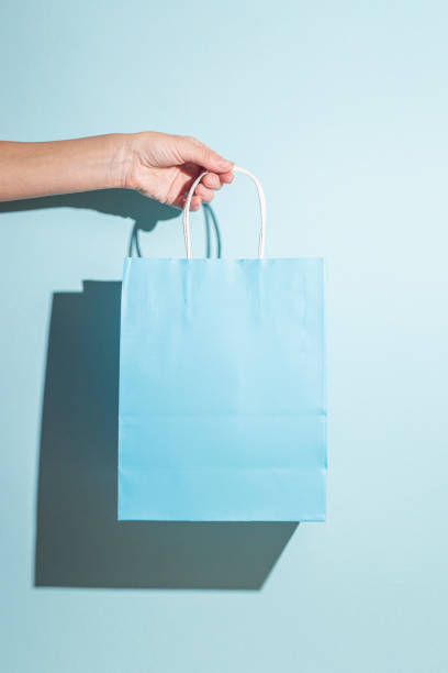istock Hand holds a paper shopping bag on a blue background. Hard shadows 1348481705
