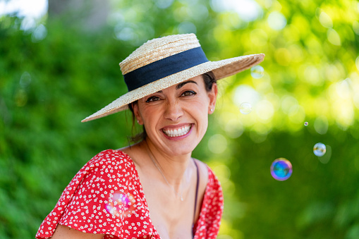 Smiling woman in red dress and straw hat having fun while playing with soap bubbles outdoors in nature. Happiness concept.