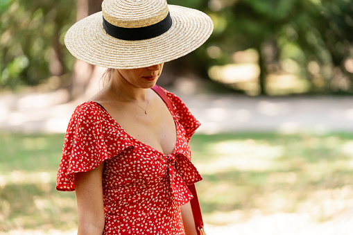 Woman in a red dress and straw hat looking down while standing outdoors in a park.