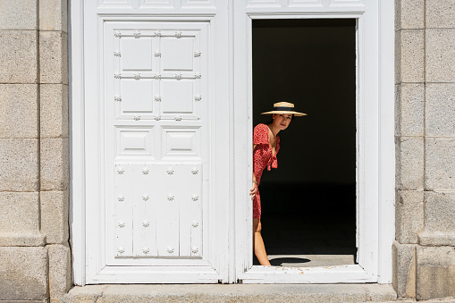 Woman in red dress and straw hat looking at the camera while hiding behind an old white wooden door. Tourism concept.