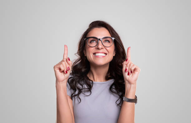 Excited woman in glasses pointing up Cheerful adult female in elegant outfit and trendy eyeglasses pointing up while standing against white background looking up stock pictures, royalty-free photos & images