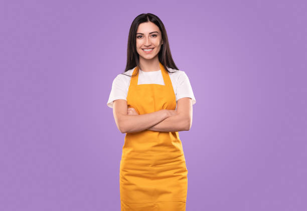 Confident cleaner looking at camera with smile Positive young woman in apron crossing arms and looking at camera with smile during housekeeping routine against violet background apron stock pictures, royalty-free photos & images