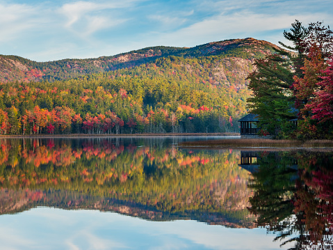 Autumn in the Adirondack region of New York and in New England boasts vivid colors because of the many tree varieties, including different shades of red, orange and yellow. This image shows these colors on the hills reflecting in calm lake water under a beautiful blue sky with wisps of clouds.