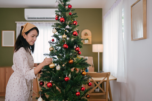 Woman decorating the christmas tree, hanging ornaments for christmas event in living room.