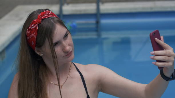 Young woman in black swimming suit and red bandana sitting on the edge of a swimming pool and taking photo of herself with cellphone. Woman enjoying summer holidays or travel vacations. stock photo