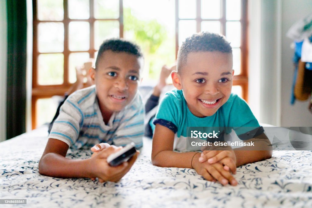 Smiling little brothers watching tv in their bedroom at home Two smiling little brothers changing channels with a remote while watching tv together on a bed at home Boys Stock Photo