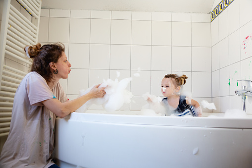 toddler boy, almost three years old, wearing bathing suit with built-in swim belt with blue and white stripes, has been finger painting in bathroom, some color is still on the wall, he is now taking a bath to wash of the color, he and his mother are blowing the foam and having a lot of fun