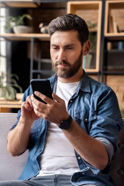 istock Caucasian man using mobile phone application sitting on sofa at home office 1348661827