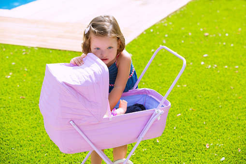 Toddler kid girl playing with baby cart in green turf grass garden
