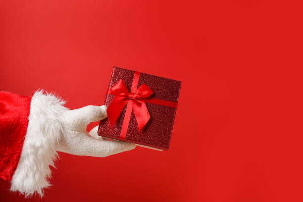 istock Santa's hand holding a gift box on red background, copy space 1348671619