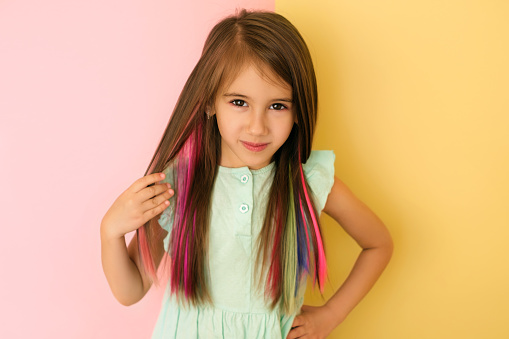Pretty cute charming girl with multicolored dyed strands of hair, isolated on a pink and yellow studio background. Safe coloring, hair care. Smooth and silky by nature. Female holds a lock of hair