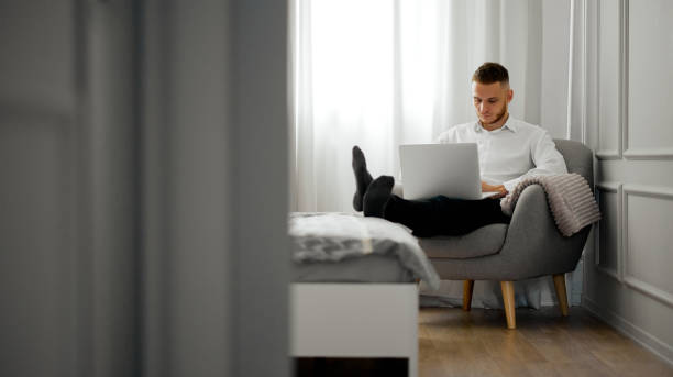 셔츠를 입은 한 청년이 침대에 다리를 대고 침실에 있는 노트북에서 일하고 있습니다. - carrying reading examining holding 뉴스 사진 이미지