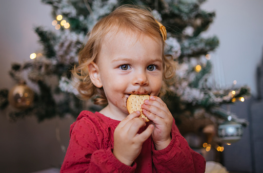 Close up photo of a little girl eating Christmas cookie in front of Christmas tree