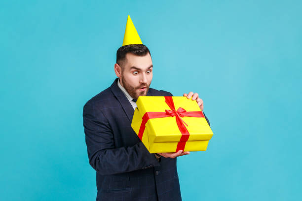 istock Businessman in official style suit with party cone on head opening gift and looking inside box, unpacking present with expression of funny amazement. 1348681132