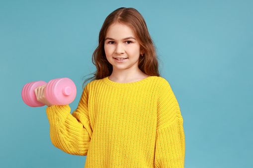 Portrait of little girl holding dumbbell in hands, expressing positive emotions, healthy lifestyle, wearing yellow casual style sweater. Indoor studio shot isolated on blue background.