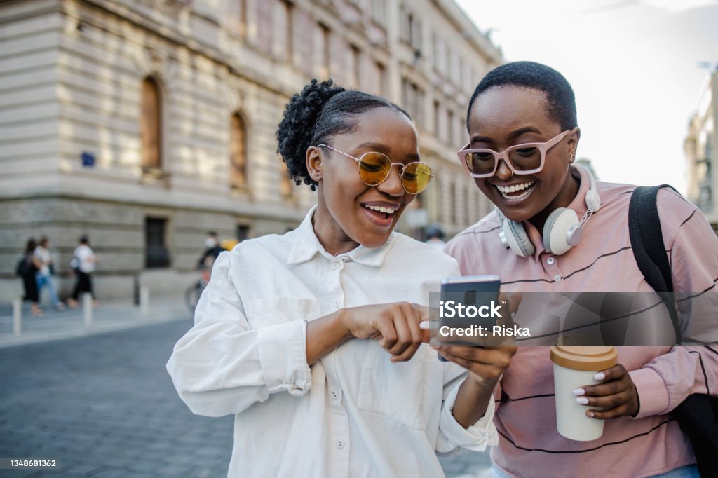 Two woman in the city, looking at the phone and smiling Two young woman standing on the street and looking at the phone Generation Z Stock Photo