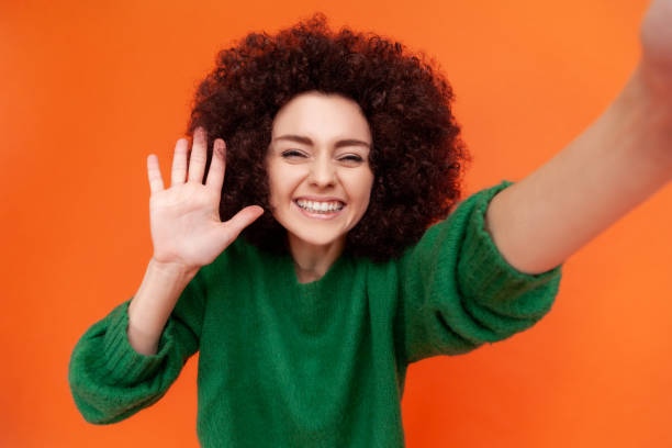 녹색 캐주얼 스타일의 스웨터를 입고 그녀의 스마트 폰으로 셀카 샷을하고 손을 흔들며 팔로워들에게 인사를 나타낸 afro 헤어 스타일에 만족한 여성. - iphone gestures 뉴스 사진 이미지