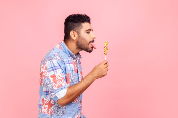 Side view of bearded handsome man wearing blue casual shirt standing tastes lollipop, dreaming licking round sweet sugary rainbow candy. Side view of bearded handsome man wearing blue casual shirt standing tastes lollipop, dreaming licking round sweet sugary rainbow candy. Indoor studio shot isolated on pink background. protruding stock pictures, royalty-free photos & images